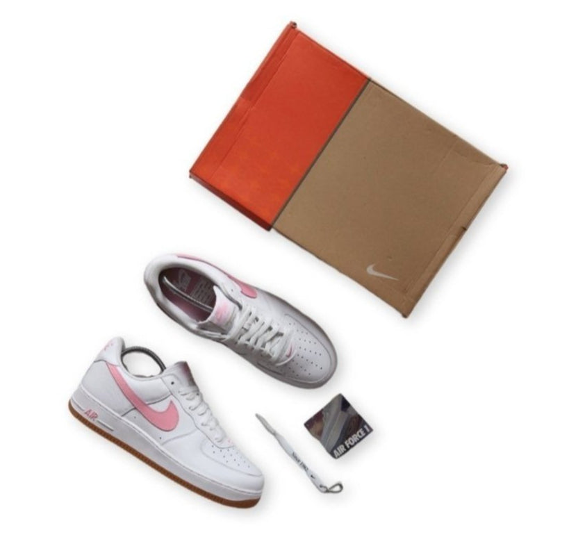 A pair of Nike Air Force 1 sneakers in white and pink, with a 25th anniversary edition tag, displayed alongside a brown shoebox, dust bag, and a toothbrush.
